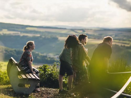 Gruppe von vier Menschen die an einem Aussichtspunkt mit Bank und Blick auf Landschaft und Tal stehen und sitzen.